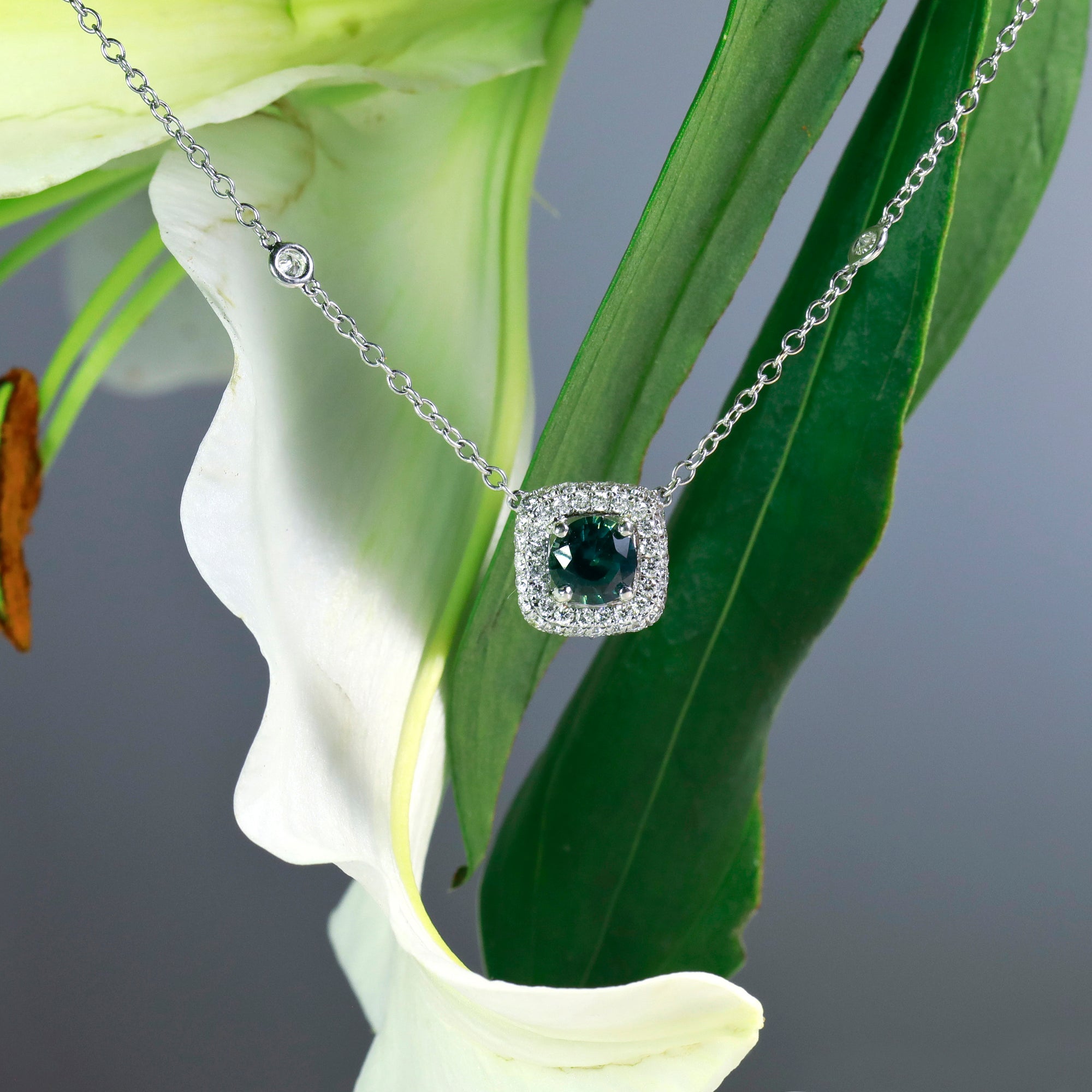 18-inch 18K white gold blue-green sapphire and diamond necklace featuring one round 1.63 carat blue-green sapphire, and round brilliant diamonds set in a cushion shaped halo and 8 bezel-set diamonds set along the chain. 